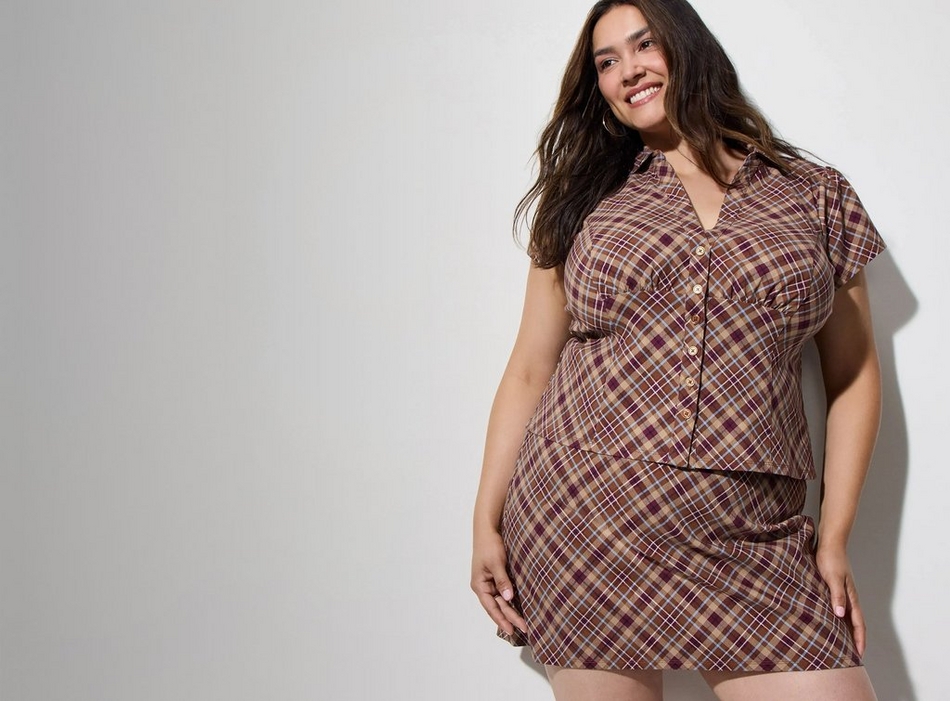 A smiling woman with long wavy dark brown hair and hoop earrings stands with one hand on her hip, wearing a brown, tan, and burgundy plaid button up short sleeve top with a collar, paired with a matching plaid mini skirt. The background is a plain light gray wall with a visible shadow.