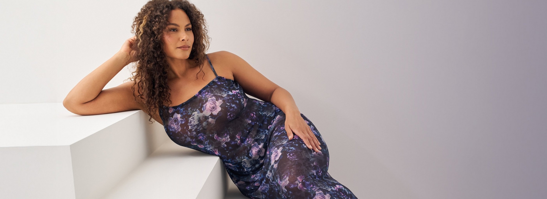 A woman with long curly brown hair reclines against a white angular platform, one hand resting near her head and the other resting on her hip. She wears a sheer navy slip dress with an allover purple and blue floral rose print, thin spaghetti straps, and a fitted bodice with a flowing skirt. The background is a plain light gray gradient wall.