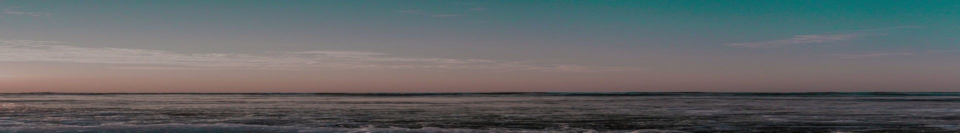 Wide panoramic photo of a beach and ocean at dusk. The sky transitions from teal at the top to pink and orange near the horizon, with wispy clouds stretching across. The dark ocean water shows small breaking waves near the horizon and rippled foreground water. No people or text appear in the image.