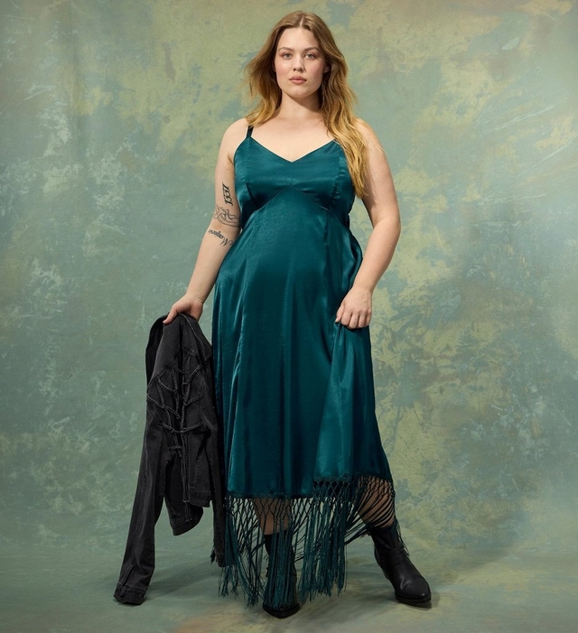 A woman with long wavy strawberry blonde hair stands holding a black denim jacket in one hand, wearing a satin teal green slip dress with a v-neckline and a fringed hem, paired with sheer patterned tights and black ankle boots. She has a small tattoo on her forearm. The background is a mottled green and gold textured surface.