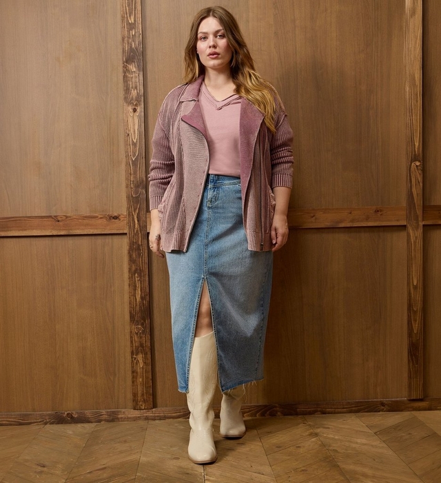 A woman with long wavy blonde and brown ombre hair stands against a wood paneled wall, wearing an open mauve pink ribbed cardigan over a matching pink v-neck top, paired with a light wash denim midi skirt featuring a front slit, and cream colored cowboy boots. She stands on a herringbone wood floor.