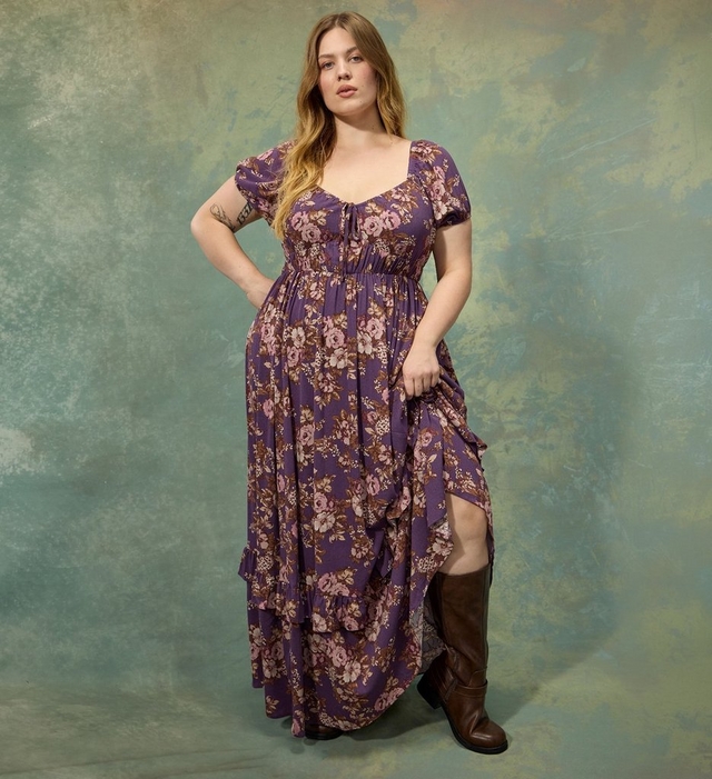 A woman with long wavy strawberry blonde hair stands with one hand on her hip, wearing a purple floral maxi dress with a deep sweetheart neckline, puff sleeves, a tie detail at the bust, and a tiered ruffled hem with a side slit, paired with brown knee high boots. She has a small tattoo on her forearm. The background is a mottled green and gold textured surface.