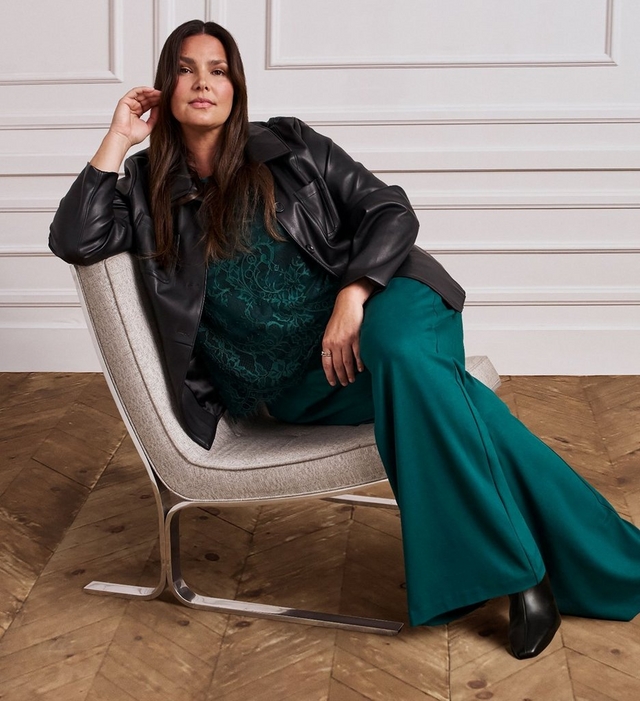 A woman with long dark hair sits sideways in a gray upholstered chair with a metal base, one hand resting near her head. She wears an open black faux leather jacket over a dark green lace top, paired with wide leg teal green trousers and black pointed toe heeled boots. The background shows white paneled walls and a herringbone wood floor.