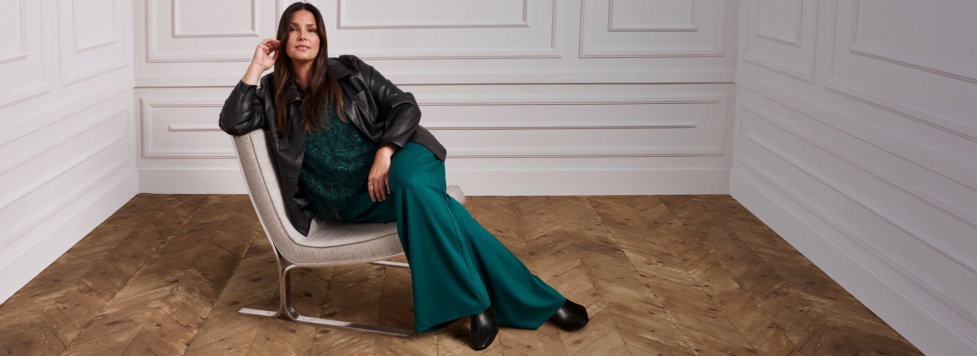 A woman with long dark hair sits sideways in a gray upholstered chair with a metal base, one hand resting near her head. She wears an open black faux leather jacket over a dark green lace top, paired with wide leg teal green trousers and black pointed toe heeled boots. The setting is a room with white paneled walls and a herringbone wood floor.