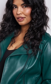 Close-up of a woman with long dark curly hair wearing a dark green faux leather moto jacket with a lapel collar and visible zipper, layered over a black top. The background is plain light gray.