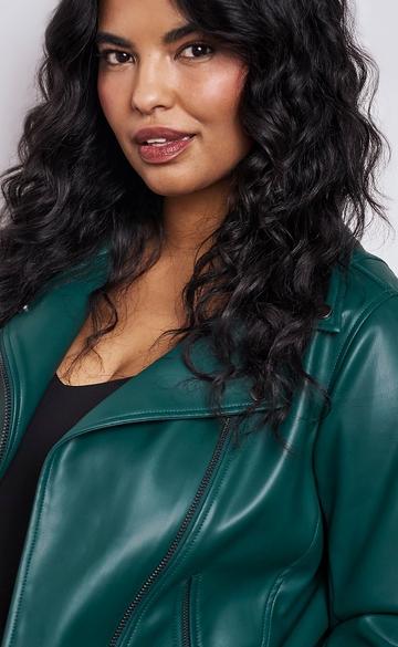 Close-up of a woman with long dark curly hair wearing a dark green faux leather moto jacket with a lapel collar and visible zipper, layered over a black top. The background is plain light gray. 