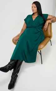 A woman with long straight dark hair sits sideways in a tan leather chair, wearing a dark green wrap style dress with short sleeves and a collar, tied at the waist, paired with black knee high heeled boots. She has visible arm tattoos and the background is plain light gray. 