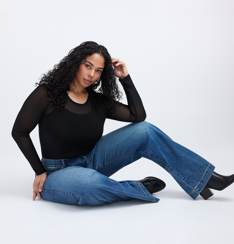 A woman with long curly dark hair sits on the floor with one hand raised near her head and the other resting on her leg. She wears a sheer black long sleeve top over a black tank layer, dark blue flared jeans, and black pointed toe ankle boots with block heels. The background is plain light gray.