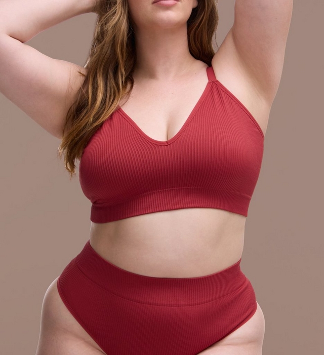 Close-up of a woman's torso with long wavy brown hair, both arms raised behind her head. She wears a ribbed dark red bralette with a V neckline and thin adjustable straps, paired with matching ribbed high waisted underwear. The background is a plain warm taupe color.