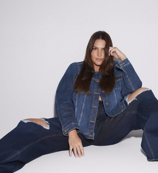 A woman with long dark brown hair sits leaning back with one hand touching her hair, wearing an open dark wash denim jacket over a matching top and dark wash wide leg jeans with distressed rips at both knees. She has a small tattoo visible on her arm and is barefoot. The background is plain white.
