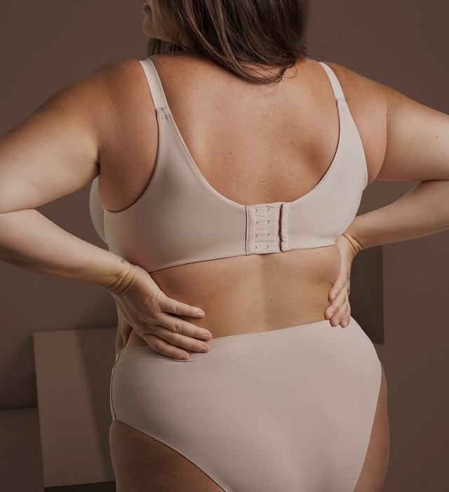 Close-up rear view of a woman with both hands resting on her lower back, wearing a nude colored bra with a hook and eye closure and adjustable straps, paired with matching nude bottoms. A small tattoo is visible on her wrist. The background is plain warm brown.