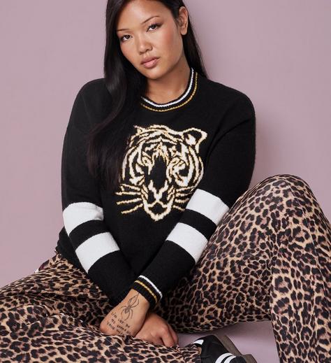 A woman with long dark hair sits with her arms resting on her knees, wearing a black crewneck sweater featuring a graphic tiger face design in gold and white with white and gold striped trim at the collar and sleeves, paired with leopard print pants. She has a small tattoo with handwritten text on her wrist. The background is plain mauve pink.