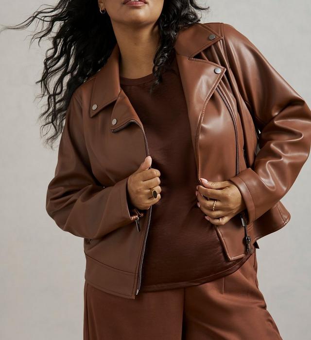 Close-up of a woman with long dark curly hair, wind blowing through it, wearing an open brown faux leather moto jacket with silver snap buttons over a brown top, paired with matching brown pants. She wears small gold hoop earrings and rings on both hands, holding the jacket open. The background is plain light gray.