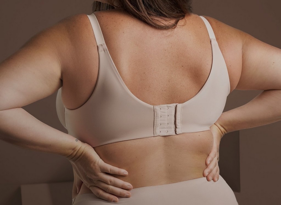 Close-up rear view of a woman with both hands resting on her lower back, wearing a nude colored bra with a hook and eye closure and adjustable straps, paired with matching nude bottoms. A small tattoo is visible on her wrist. The background is plain warm brown.
