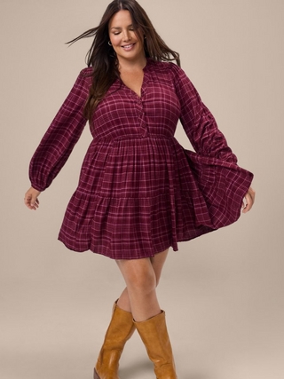 A woman with long dark wavy hair twirls with her arms out, wearing a burgundy and pink plaid long sleeve fit and flare dress with a button placket and tiered skirt, paired with tan suede knee high boots. The background is plain light beige. 