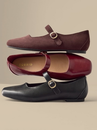 Three pairs of mary jane style flats stacked and arranged against a plain beige background, in burgundy suede, dark red patent leather with a visible 'TORRID' brand label on the insole, and black leather, each featuring a rounded toe and a buckled strap.