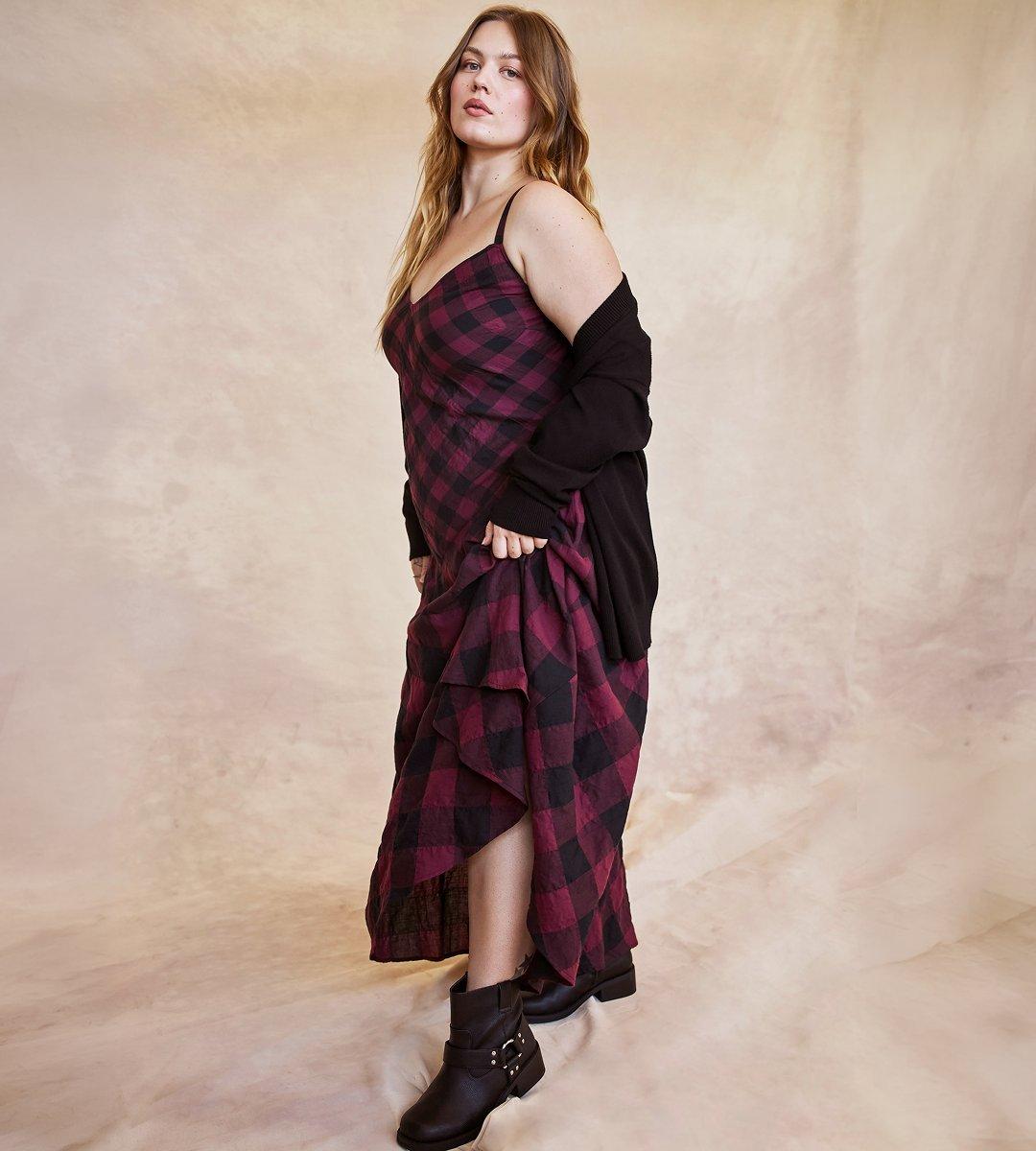 A model with long wavy strawberry-blonde hair stands in three-quarter profile on a mottled peach and cream painted backdrop, body turned away and face toward the camera. She wears a burgundy and black buffalo check slip dress with thin spaghetti straps and a V neckline, worn under a black knit cardigan that has slipped off both shoulders and hangs at her upper arms. Her right hand gathers a handful of the long check skirt, lifting it to one side so the hem sweeps into a diagonal drape and reveals a darker under-layer. On her feet are dark brown leather ankle boots with a harness strap and small silver studs. The floor is covered in the same soft cream fabric as the backdrop.