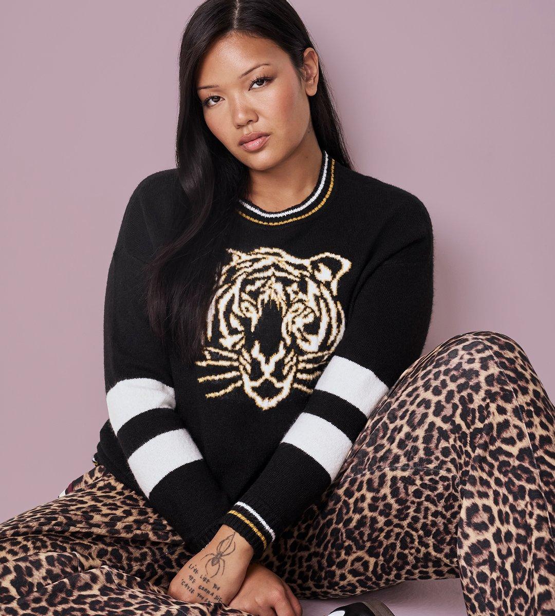 A model with long straight black hair sits on the floor against a dusty mauve backdrop, knees drawn up and forearms crossed over them, looking directly at the camera. She wears an oversized black knit sweater with a large tiger head knitted into the chest in cream and gold, a ribbed crewneck collar striped in white and gold, and sleeves banded with wide black and white stripes and gold-trimmed cuffs. Her pants are a tan and black leopard print. Fine-line tattoos, including a spider and small script lettering, are visible on her forearm and wrist. The toe of a black sneaker shows at the lower edge of the frame.