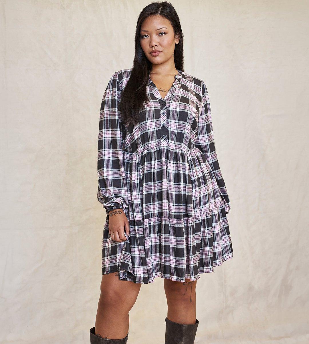 A model with long straight black hair stands facing the camera on a cream canvas backdrop with soft creases and shadows. She wears a tiered babydoll mini dress in black, white, and pink plaid, with a band collar, a short button placket at the chest, a gathered seam under the bust, and long blouson sleeves ending in buttoned cuffs. A thin gold script nameplate necklace sits at her collarbone, and she wears a ring on her right hand. Fine-line tattoos are visible on her right wrist and on her leg below the hem. She wears tall dark brown leather boots that reach the knee.