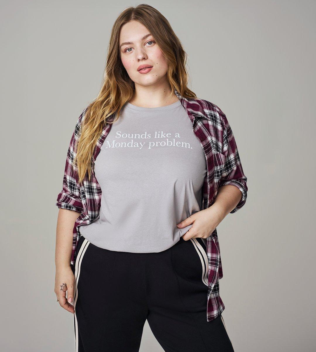 A model with long wavy blonde hair stands facing the camera against a plain grey studio backdrop, one hand tugging the hem of her shirt. She wears a light grey crewneck T-shirt printed across the chest in white serif type with the words 'Sounds like a Monday problem.' Over it hangs an open plaid flannel shirt in burgundy, white, and black with the sleeves rolled to the elbow. Her black track pants have a cream stripe down the side seam. A small fine-line tattoo sits on her left forearm and she wears a thin ring on one finger.