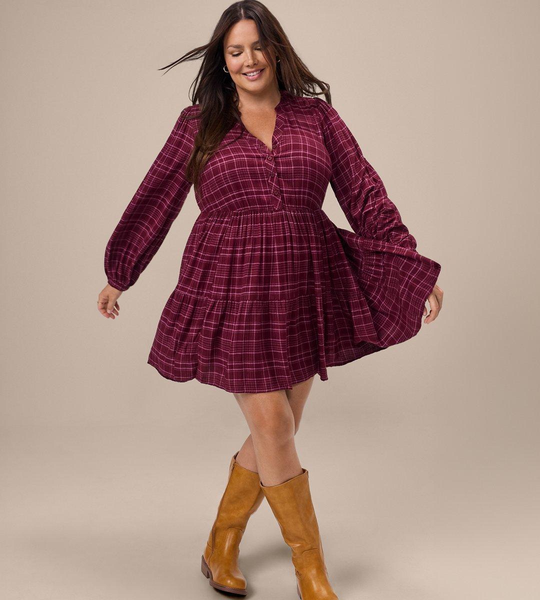 A model with long brown hair stands mid-twirl on a warm beige backdrop, eyes closed and smiling, arms loose at her sides as her hair and skirt swing outward. She wears a burgundy plaid tiered babydoll mini dress with fine pink and white grid lines, a band collar, a short button placket, a gathered waist seam, and full blouson sleeves with elasticated cuffs. The skirt lifts and flares to her left with the movement. She wears tall camel-brown leather boots that reach just below the knee, and small gold hoop earrings.