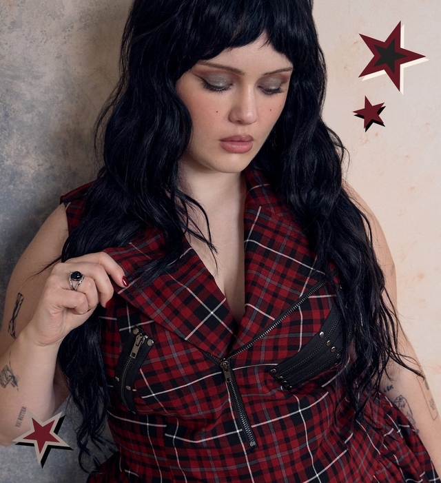 A woman with long dark wavy hair and bangs looks down, wearing a sleeveless red and black plaid top with buckle and zipper details and a deep v-neckline, with visible tattoos on her arm and a ring on her hand. Red star graphics are scattered across the plain beige background.