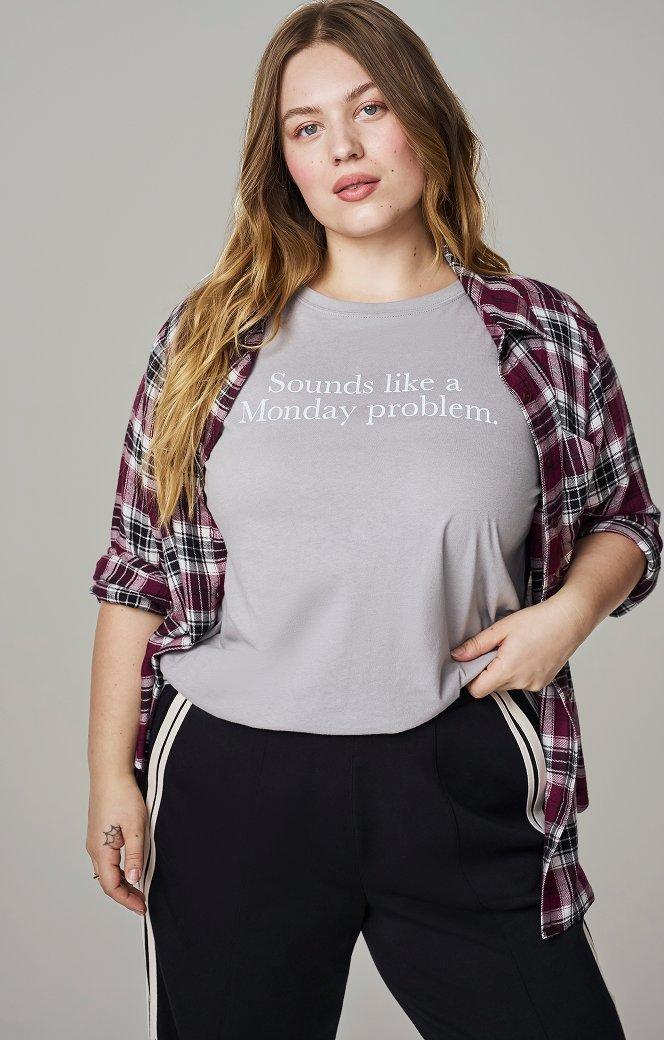 A model with long wavy blonde hair stands facing the camera against a plain grey backdrop, one hand resting near her hip. She wears a light grey crewneck T-shirt printed across the chest in white serif type with the words 'Sounds like a Monday problem.' Over it hangs an open plaid flannel shirt in burgundy, white, and black. Her black track pants have a cream stripe down the side seam.