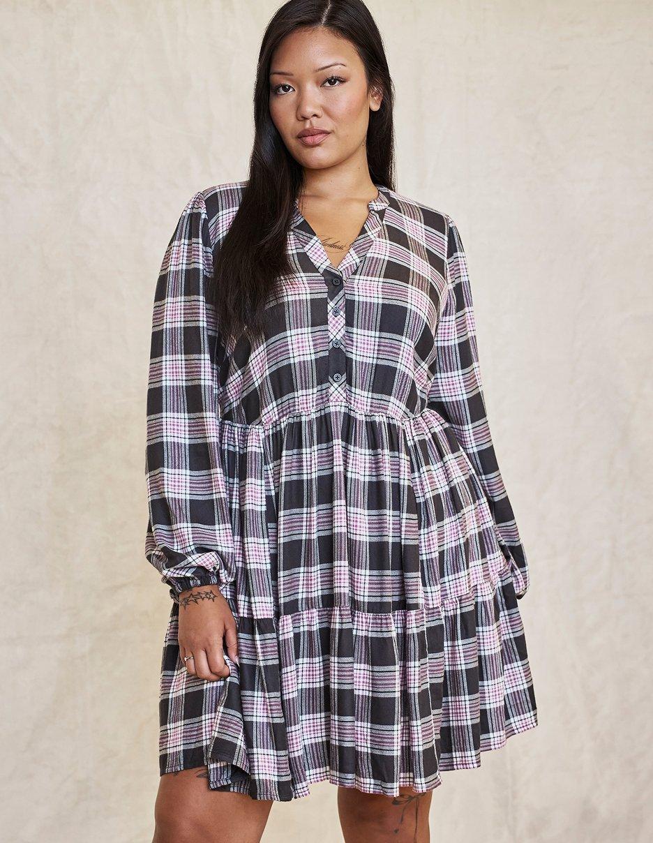 A model with long straight black hair stands facing the camera on a cream canvas backdrop. She wears a tiered babydoll mini dress in black, white, and pink plaid with a band collar, a short button placket at the chest, and long blouson sleeves with buttoned cuffs. A thin gold necklace sits at her collarbone and a small tattoo is visible on her wrist.