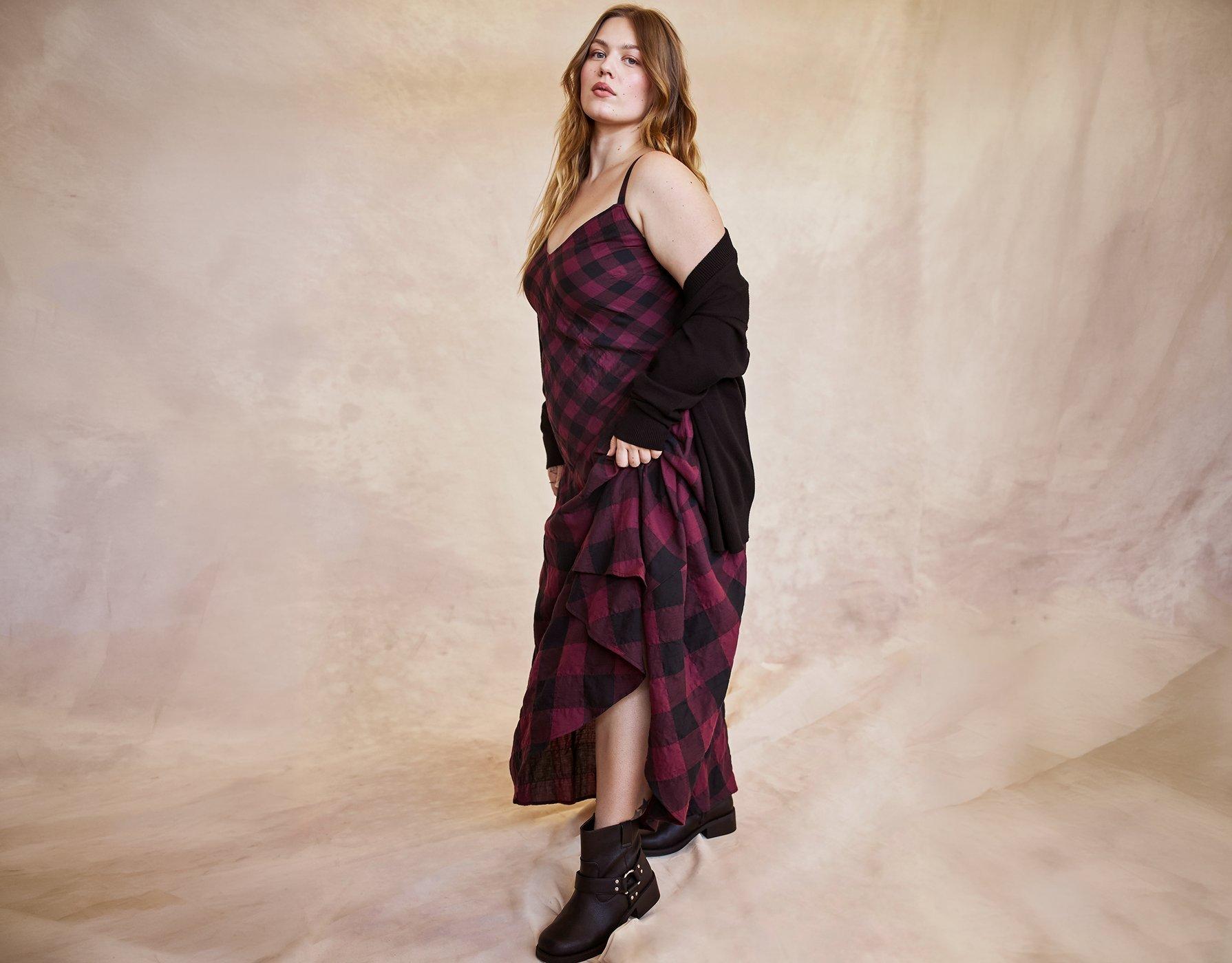 A model with long wavy strawberry blonde hair stands in three-quarter profile on a mottled peach and cream painted backdrop, looking toward the camera. She wears a burgundy and black buffalo check slip dress with thin straps and a black knit cardigan slipped off her shoulders. One hand gathers the long skirt to the side, lifting the hem into a diagonal drape. She wears dark brown leather ankle boots with a harness strap and small studs.