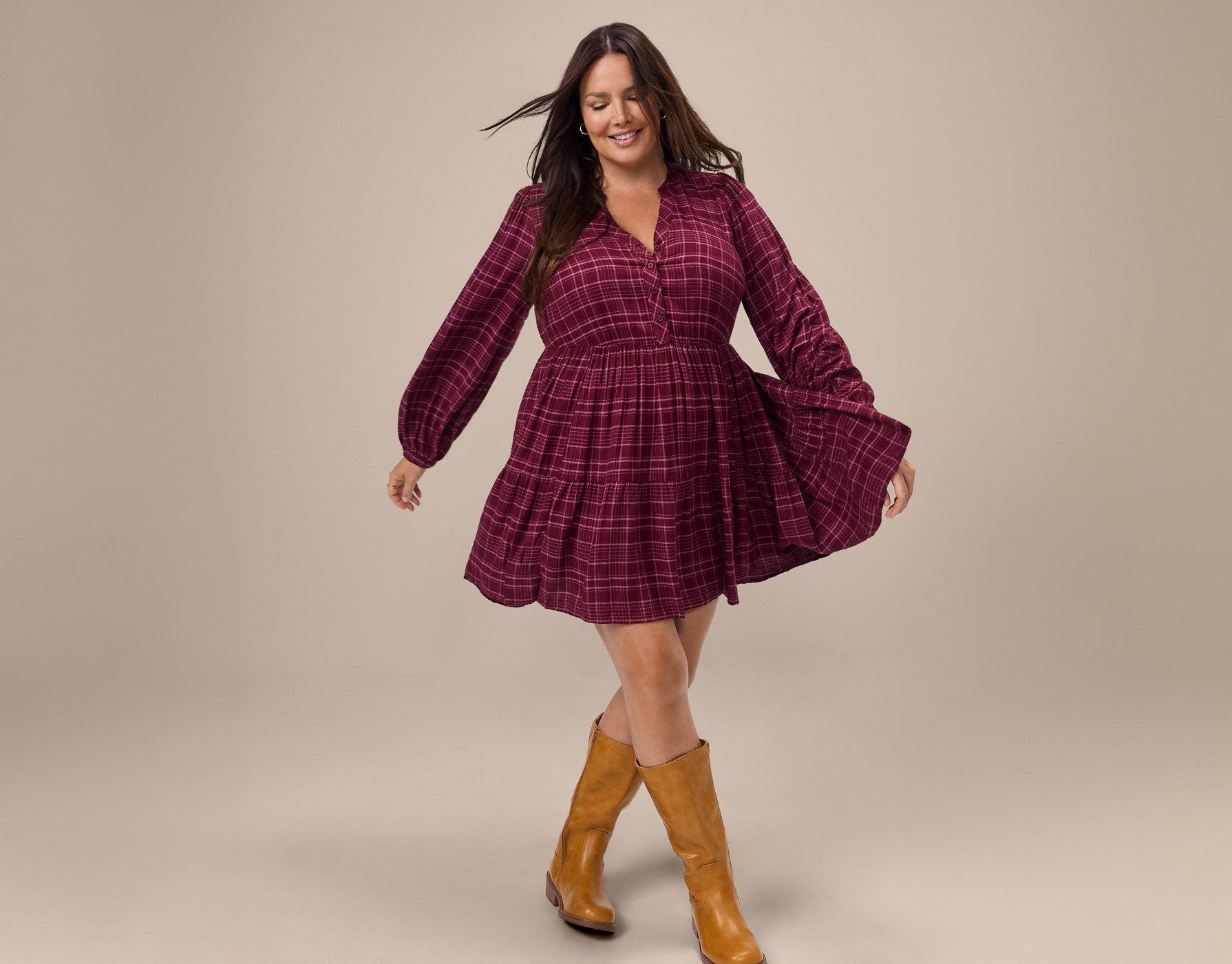 A model with long dark hair is caught mid-twirl against a plain warm taupe backdrop, smiling as her hair swings behind her. She wears a burgundy plaid tiered babydoll mini dress with fine pink grid lines, a band collar, a short button placket, and full blouson sleeves. The skirt flares out to one side with the movement. She wears tall camel-brown leather boots that reach just below the knee.