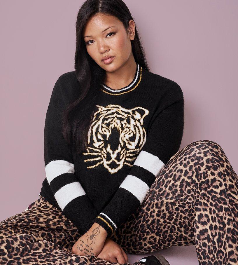 A model with long straight black hair sits against a dusty mauve backdrop, knees drawn up and arms crossed over them, looking directly at the camera. She wears a black knit sweater with a large cream and gold tiger head on the chest, a striped ribbed collar, and wide black and white stripes on the sleeves. Her pants are a tan and black leopard print. A fine-line tattoo is visible on her wrist.