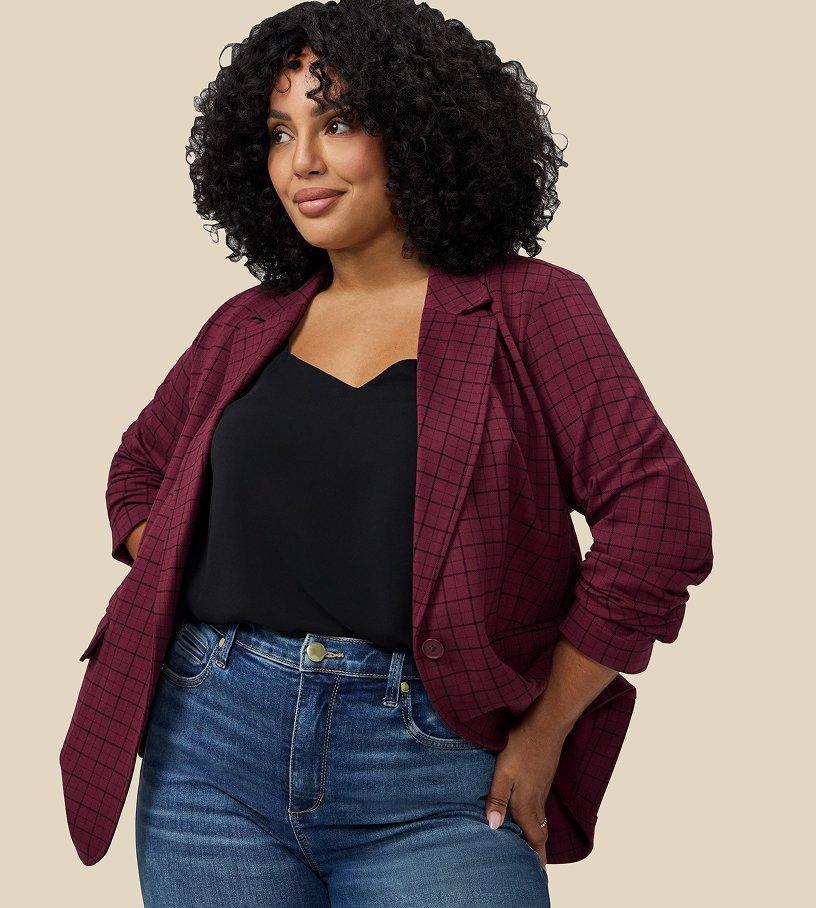 A model with voluminous dark curly hair stands against a plain beige backdrop, looking off to one side with a slight smile, one hand tucked into her jeans pocket. She wears an open burgundy blazer with a black windowpane check and sleeves pushed up the forearm, over a black V-neck camisole, with medium wash blue jeans.