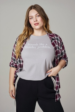 A woman with long wavy strawberry blonde hair stands with one hand near her waist, wearing an open burgundy, black, and white plaid button-up shirt over a light gray t-shirt with white text reading 'Sounds like a Monday problem.', paired with black pants featuring a white and burgundy side stripe. The background is plain light gray.