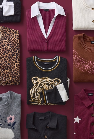 Overhead flat lay of folded clothing items arranged in a grid against a dark maroon background. Items include a leopard print cardigan, a burgundy button-up vest layered over a white collared shirt, a cream cable knit sweater, a black sweater featuring a tiger graphic with folded white sleeves, a brown sweater with paisley print detailing, a gray sweater with an embroidered floral design, a black button-up shirt, and a burgundy sweater with a white star graphic. No people or text appear in the image. 