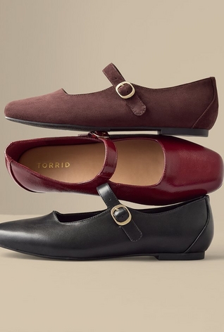 Three pairs of mary jane style flats stacked and arranged against a plain beige background, in burgundy suede, dark red patent leather with a visible 'TORRID' brand label on the insole, and black leather, each featuring a rounded toe and a buckled strap. 