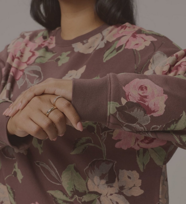 A close-up of a model from the chin to the waist against a light grey background, with ends of dark hair falling at the top of the frame. She wears a loose dusty plum crewneck sweatshirt printed with large pink and rose roses and sage green leaves, with ribbed cuffs. Her left arm is bent across her body with her right hand resting over the wrist, showing thin gold rings on her fingers and light nude nails.
