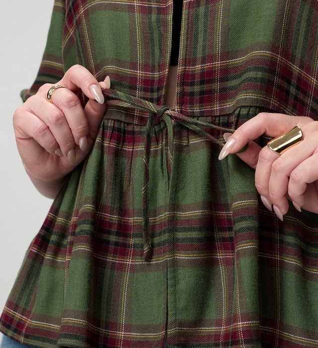 A close-up of a model's midsection against a light grey background, showing a loose olive green plaid top crossed with burgundy, cream, and golden tan stripes. Both hands hold the thin self-fabric drawstring ties at the waist, pulling them into a loose bow that gathers the fabric into a flared peplum below. A dark layer shows through the open front above the waist. The model has pale pink almond-shaped nails, a thin ring on her left hand, and a wide gold ring on her right hand.