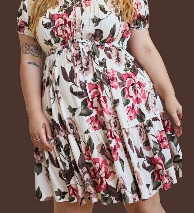 A model shown from the shoulders to mid-thigh against a dark chocolate brown background, with ends of long blonde hair falling at the top of the frame. She wears a short cream dress printed with large pink and rose roses and dark green and brown leaves. The dress has short puffed sleeves, a fitted bodice with a small ruffled front placket, a gathered tie waist with thin drawstring ends hanging down the front, and a full tiered skirt ending above the knee. Tattoos are visible on her right upper arm and inner forearm, and both arms hang relaxed at her sides.