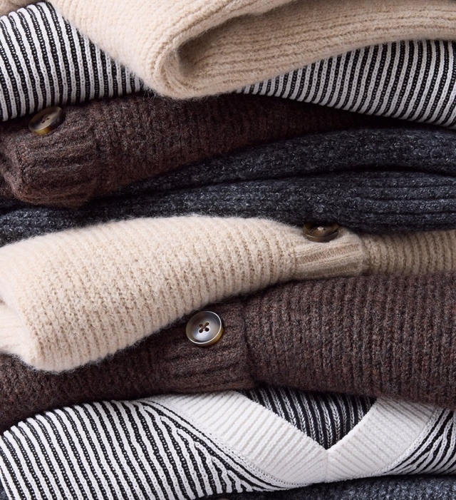 A close-up of a model's midsection against a light grey background, showing a loose olive green plaid top crossed with burgundy, cream, and golden tan stripes. Both hands hold the thin self-fabric drawstring ties at the waist, pulling them into a loose bow that gathers the fabric into a flared peplum below. A dark layer shows through the open front above the waist. The model has pale pink almond-shaped nails, a thin ring on her left hand, and a wide gold ring on her right hand.