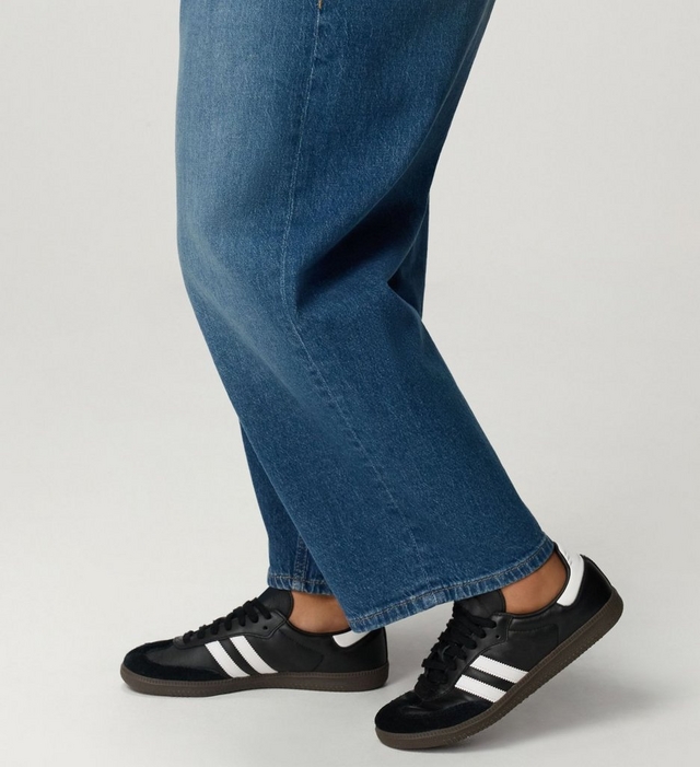 A close-up of a model's lower legs from the knee down, shown in profile mid-step against a plain light grey background. She wears medium-wash blue wide-leg jeans with visible fading along the front crease, a center seam running down the leg, and a stitched hem that falls just above the ankle. On her feet are low-profile black leather sneakers with black suede toe overlays, three white diagonal side stripes, black laces, white heel tabs, and dark gum rubber soles.