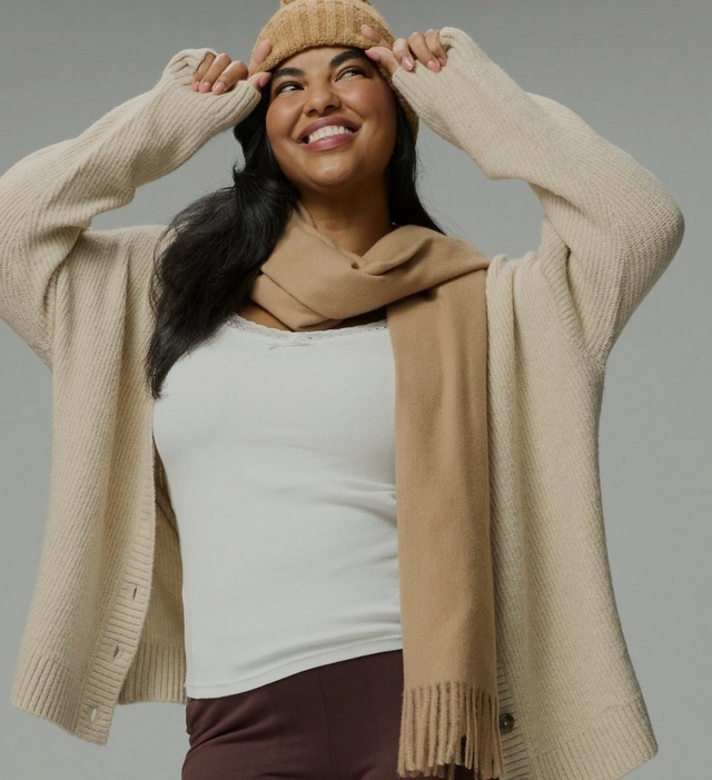 A model with long dark hair smiles broadly and looks up to her left against a plain grey background, both hands raised to the brim of a camel ribbed knit beanie. She wears an oversized oatmeal ribbed knit cardigan worn open, with dropped shoulders, long sleeves pushed up her wrists, ribbed cuffs and hem, and dark buttons along the front edge. Underneath is a fitted ivory ribbed tank with a lace-trimmed scoop neckline and a small bow at the center. A long camel scarf with fringed ends is looped once around her neck, with one end hanging down the front. The top of dark chocolate brown pants shows at the bottom of the frame.