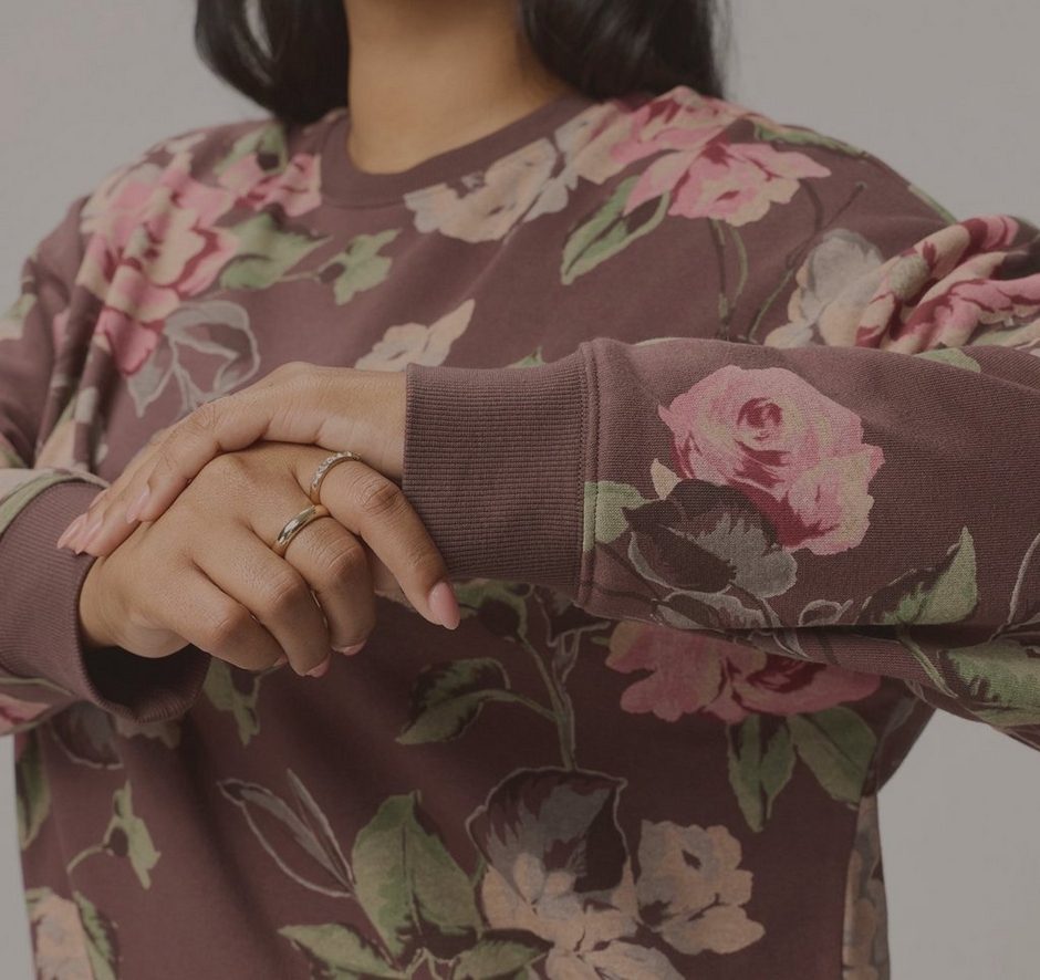 A close-up of a model from the chin to the waist against a light grey background, with ends of dark hair falling at the top of the frame. She wears a loose dusty plum crewneck sweatshirt printed with large pink and rose roses and sage green leaves, with ribbed cuffs. Her left arm is bent across her body with her right hand resting over the wrist, showing thin gold rings on her fingers and light nude nails.