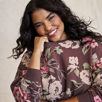 A model shown from the waist up against a light cream background, with long, dark, curly hair falling past her shoulders. She smiles broadly at the camera with her chin resting on her right hand. She wears a loose dusty plum sweatshirt printed with large pink and cream roses and sage green leaves, with ribbed cuffs.