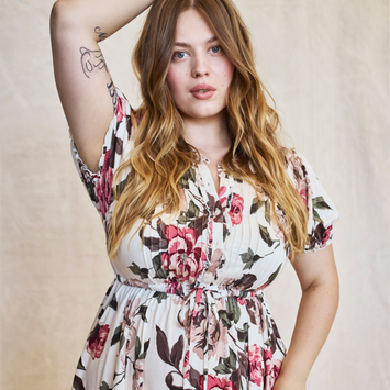 A model shown from the head to the hips against a textured cream backdrop, with long, wavy, light brown hair falling past her chest. She looks at the camera with a soft expression, her right arm raised behind her head showing tattoos on her inner upper arm and forearm. She wears a cream dress printed with large pink and red roses and dark green leaves, with short flutter sleeves, a buttoned front placket, and a gathered drawstring waist.