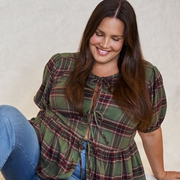 A model seated against a light cream background, with long, straight, dark brown hair falling over her shoulders. She smiles and looks down, one knee bent and her left hand resting on the floor beside her. She wears a green plaid top crossed with red, cream, and dark brown stripes, with short puffed sleeves, small tie closures down the front, and a gathered peplum, paired with light blue jeans.