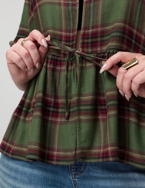 A close-up of a model's midsection against a light grey background, showing a loose olive green plaid top crossed with burgundy, cream, and golden tan stripes. Both hands hold the thin self-fabric drawstring ties at the waist, pulling them into a loose bow that gathers the fabric into a flared peplum below. A dark layer shows through the open front above the waist. The model has pale pink almond-shaped nails, a thin ring on her left hand, and a wide gold ring on her right hand.