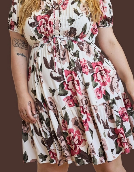 A model shown from the shoulders to mid-thigh against a dark chocolate brown background, with ends of long blonde hair falling at the top of the frame. She wears a short cream dress printed with large pink and rose roses and dark green and brown leaves. The dress has short puffed sleeves, a fitted bodice with a small ruffled front placket, a gathered tie waist with thin drawstring ends hanging down the front, and a full tiered skirt ending above the knee. Tattoos are visible on her right upper arm and inner forearm, and both arms hang relaxed at her sides.