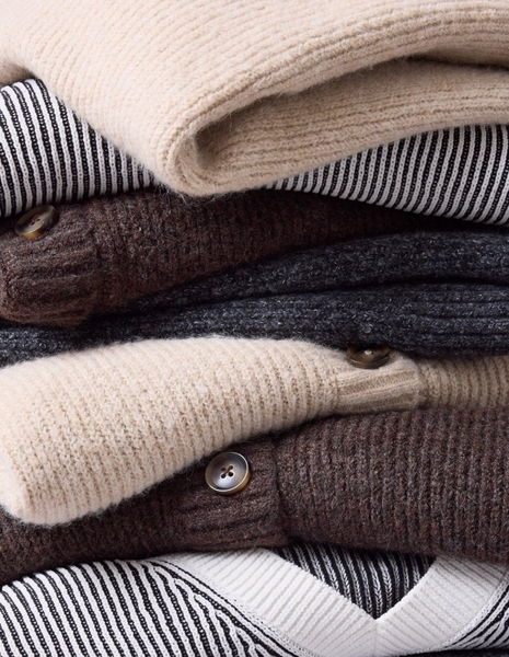 A close-up of a model's midsection against a light grey background, showing a loose olive green plaid top crossed with burgundy, cream, and golden tan stripes. Both hands hold the thin self-fabric drawstring ties at the waist, pulling them into a loose bow that gathers the fabric into a flared peplum below. A dark layer shows through the open front above the waist. The model has pale pink almond-shaped nails, a thin ring on her left hand, and a wide gold ring on her right hand.