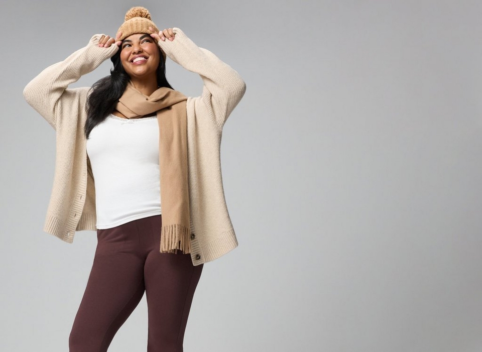 A model with long dark hair smiles broadly and looks up to her left against a plain grey background, both hands raised to the brim of a camel ribbed knit beanie. She wears an oversized oatmeal ribbed knit cardigan worn open, with dropped shoulders, long sleeves pushed up her wrists, ribbed cuffs and hem, and dark buttons along the front edge. Underneath is a fitted ivory ribbed tank with a lace-trimmed scoop neckline and a small bow at the center. A long camel scarf with fringed ends is looped once around her neck, with one end hanging down the front. The top of dark chocolate brown pants shows at the bottom of the frame.