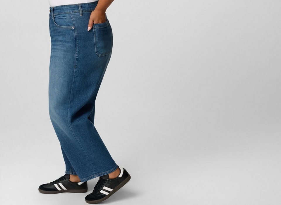A close-up of a model's lower legs from the knee down, shown in profile mid-step against a plain light grey background. She wears medium-wash blue wide-leg jeans with visible fading along the front crease, a center seam running down the leg, and a stitched hem that falls just above the ankle. On her feet are low-profile black leather sneakers with black suede toe overlays, three white diagonal side stripes, black laces, white heel tabs, and dark gum rubber soles.